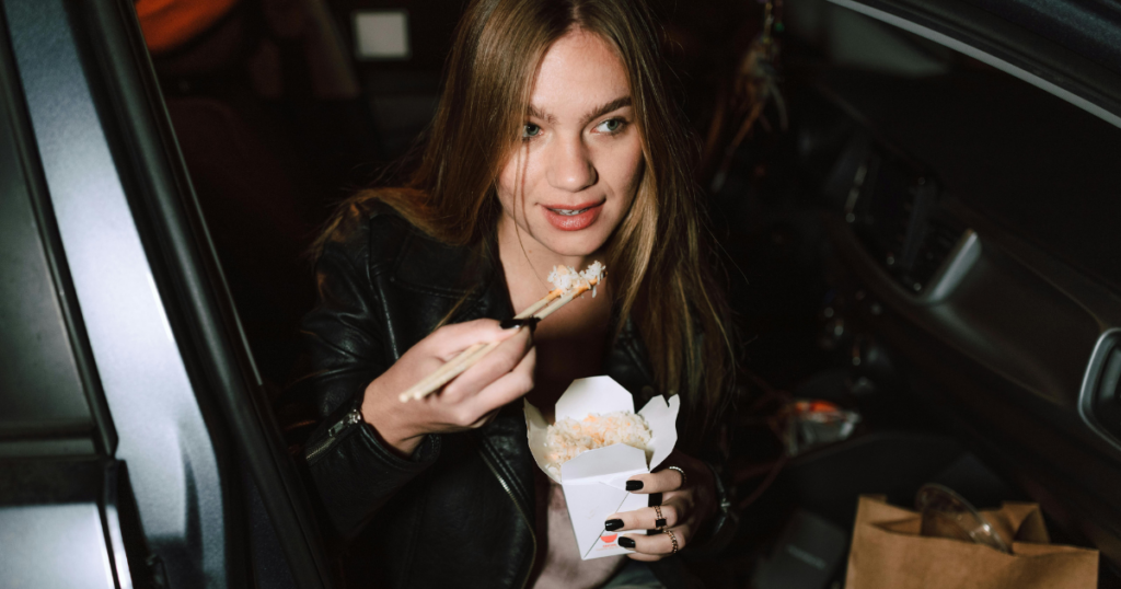 image - My Teen Mental Health Woman in a leather jacket sits in a car, holding a takeout box of sushi and chopsticks ready to eat at night.