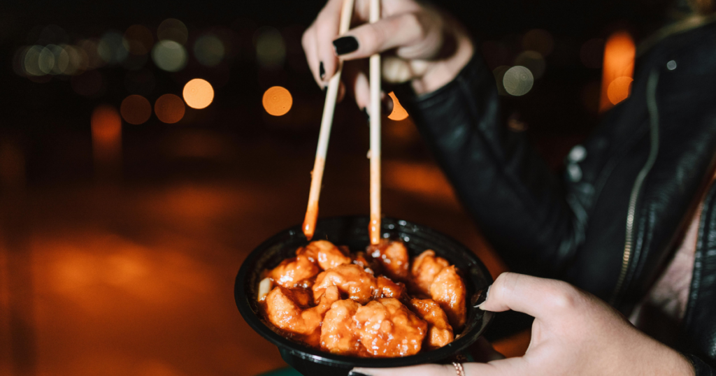 image - My Teen Mental Health Close-up of a person with a black leather jacket holding a bowl of orange glaze-coated fried bites while using chopsticks at night; bokeh lights in the background.