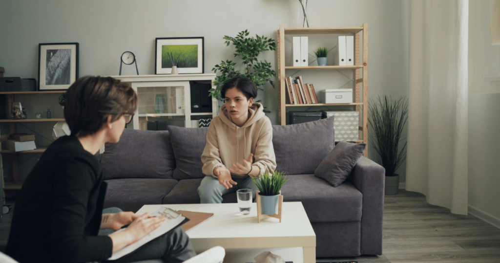 image - My Teen Mental Health Two people converse in a cozy living room; a person on a gray sofa gestures while the other takes notes at a white coffee table.