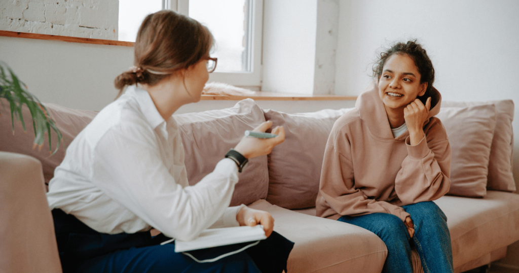 image - My Teen Mental Health Therapist sits with notebook and pen, talking with a young woman in a pink hoodie on a beige couch in a bright room.