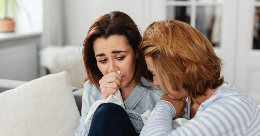 image - My Teen Mental Health Two women sit close on a sofa; one sneezes or blows her nose with a tissue while the other offers support.