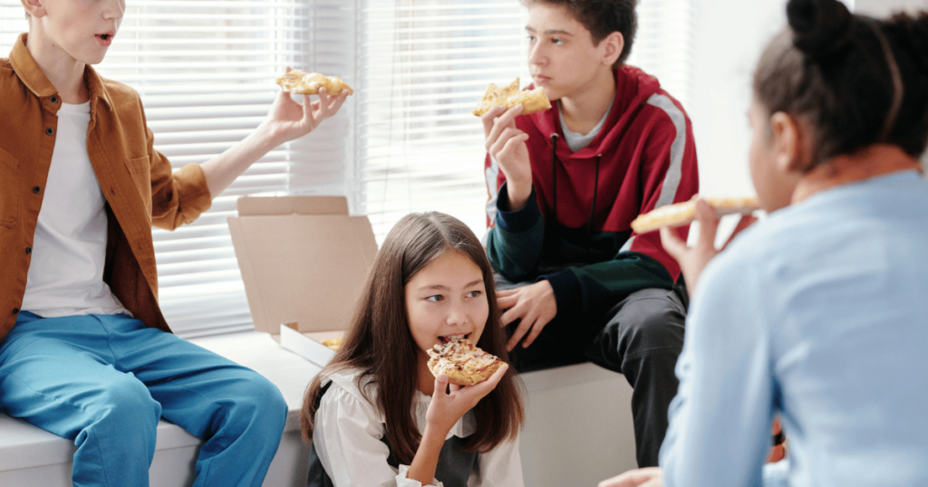 image - My Teen Mental Health Four teens sit by a window, sharing pizza and chatting in a bright room.
