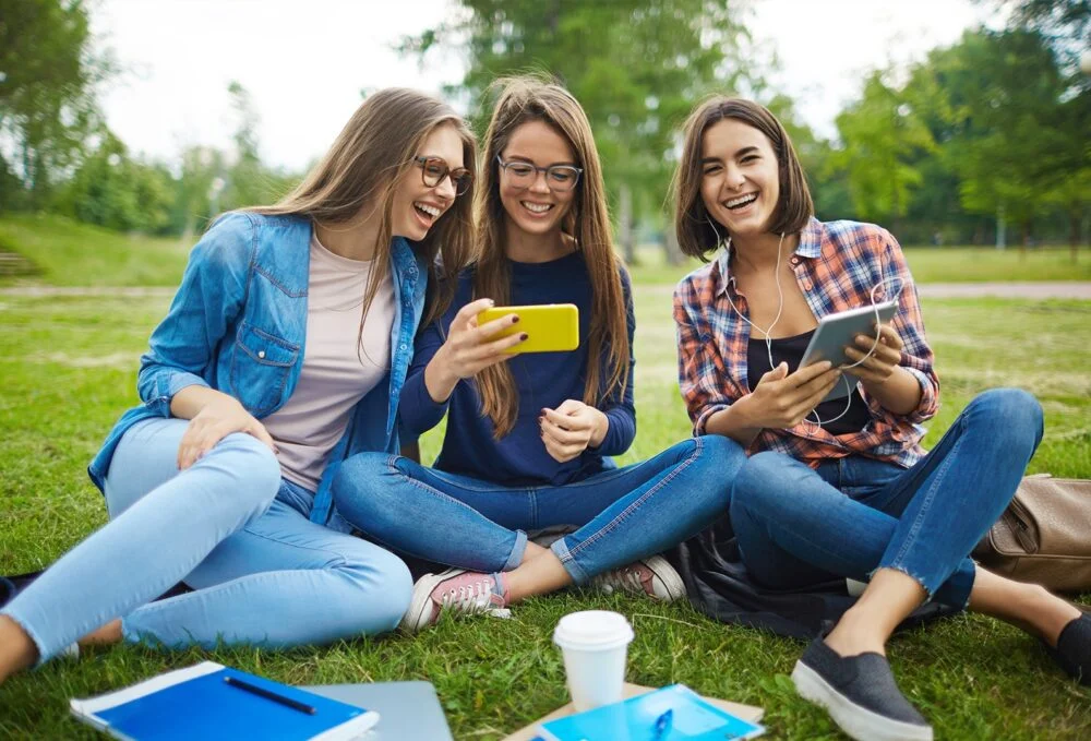 Three friends sit on the grass in a park, laughing while using a yellow smartphone and a tablet nearby.