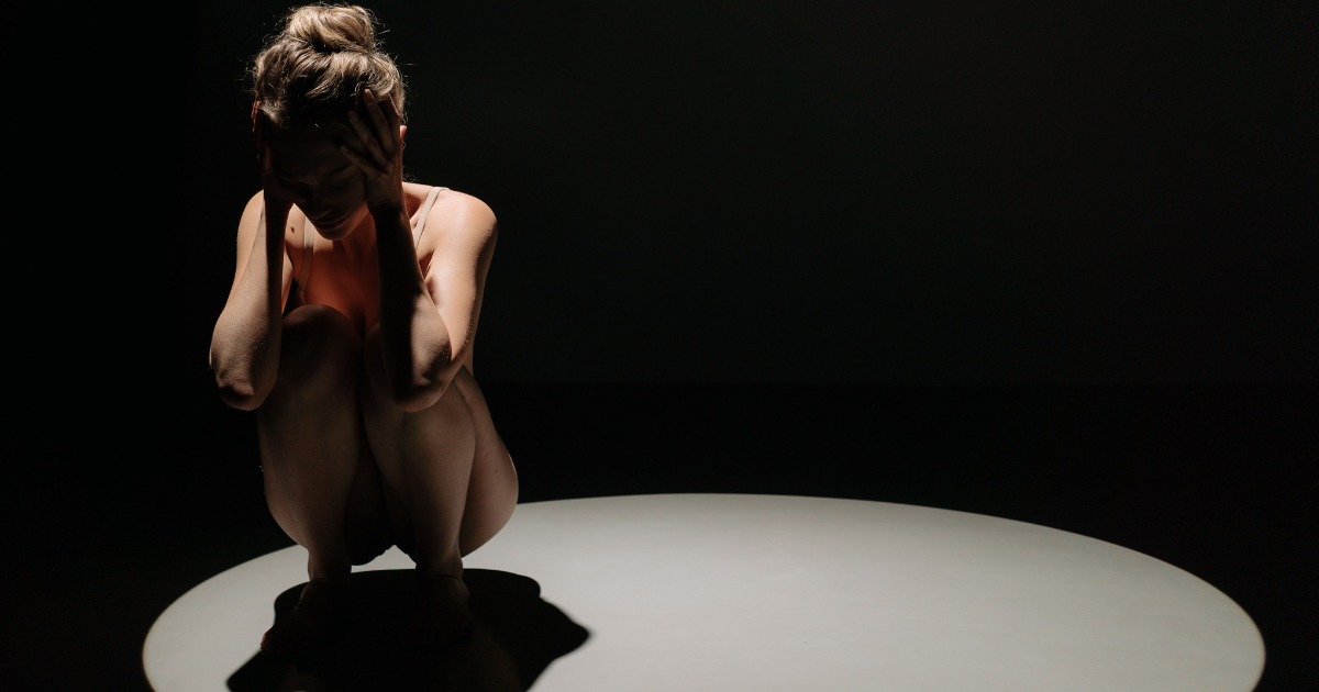 A person sits curled up on a lit circular spotlight, head in hands, in a dark room, expressing distress or isolation.