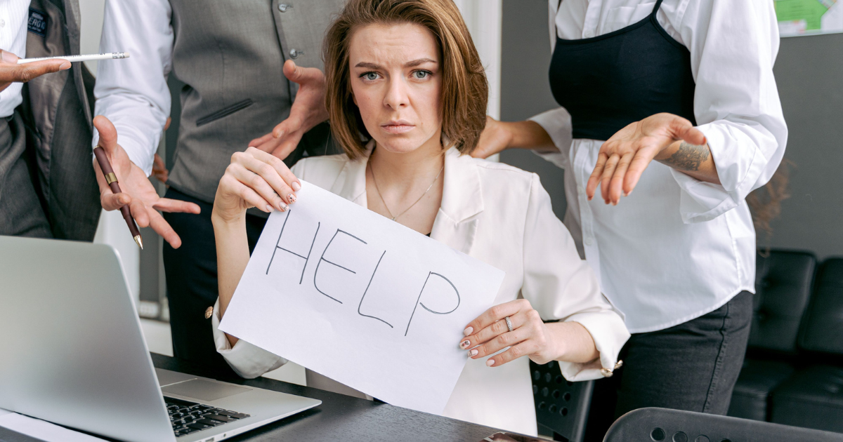 Woman in a white blazer sits at a desk with a laptop, holding a handwritten sign that reads HELP while colleagues crowd around her, gesturing for attention.