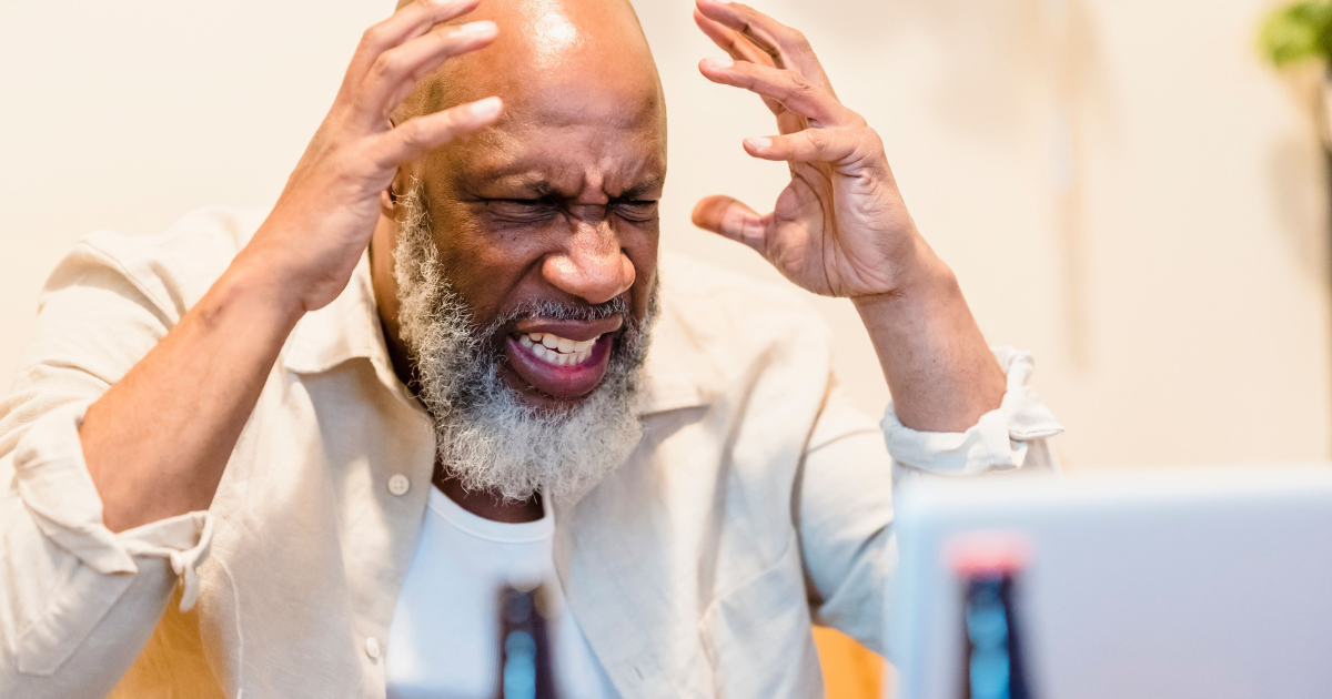 Middle-aged man with a gray beard grimaces in frustration, hands on his head while looking at a laptop.