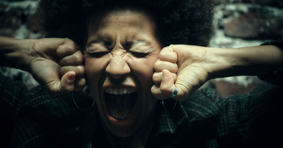 Woman with curly hair covering ears, mouth wide in a scream.