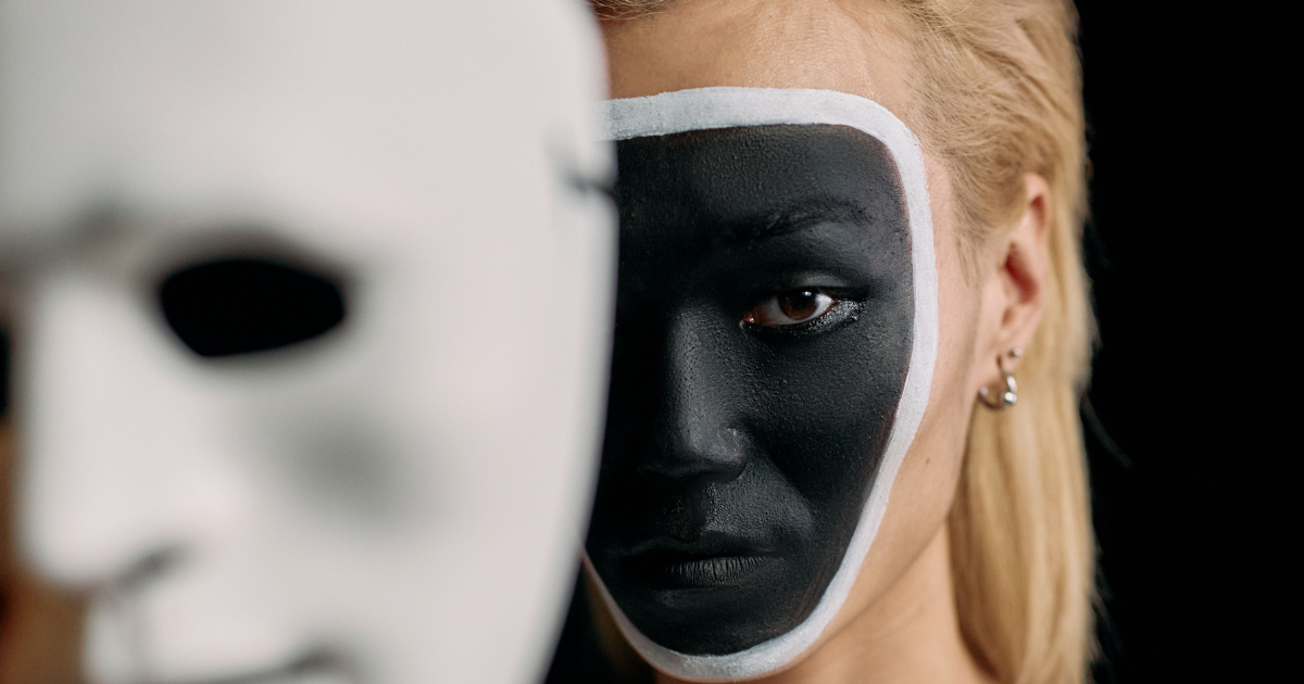 Close-up of a person with black face paint outlined in white around the face, looking toward the camera, with a blurred white mask in the foreground.
