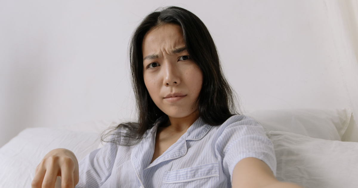 Woman in pale striped pajamas sits on a bed, looking puzzled at the camera.